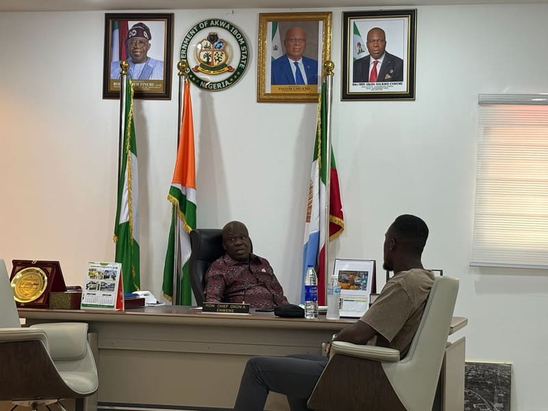 Pr. Joseph Ante seated across the desk from the Executive Chairman in his office, with flags and official portraits on the wall behind the desk
