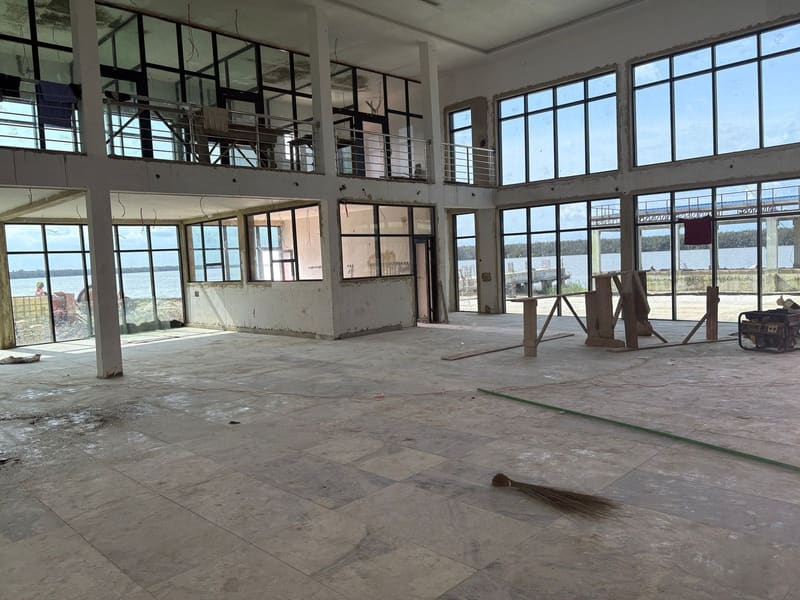 A large empty hall with tall window frames and an unfinished floor, overlooking the water